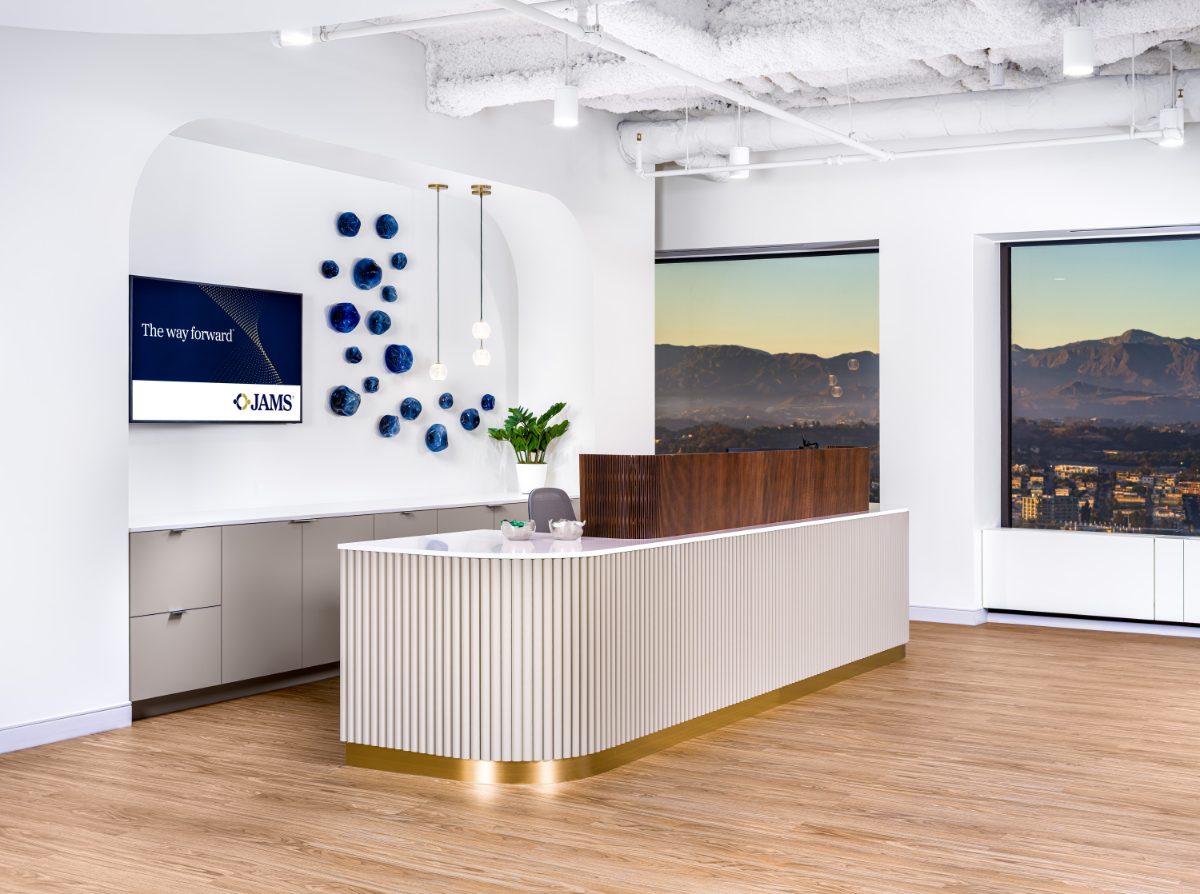 Close-up reception desk at JAMS Los Angeles — fluted cream millwork with brass base, walnut divider, blue ceramic art, and San Gabriel Mountains at golden hour.