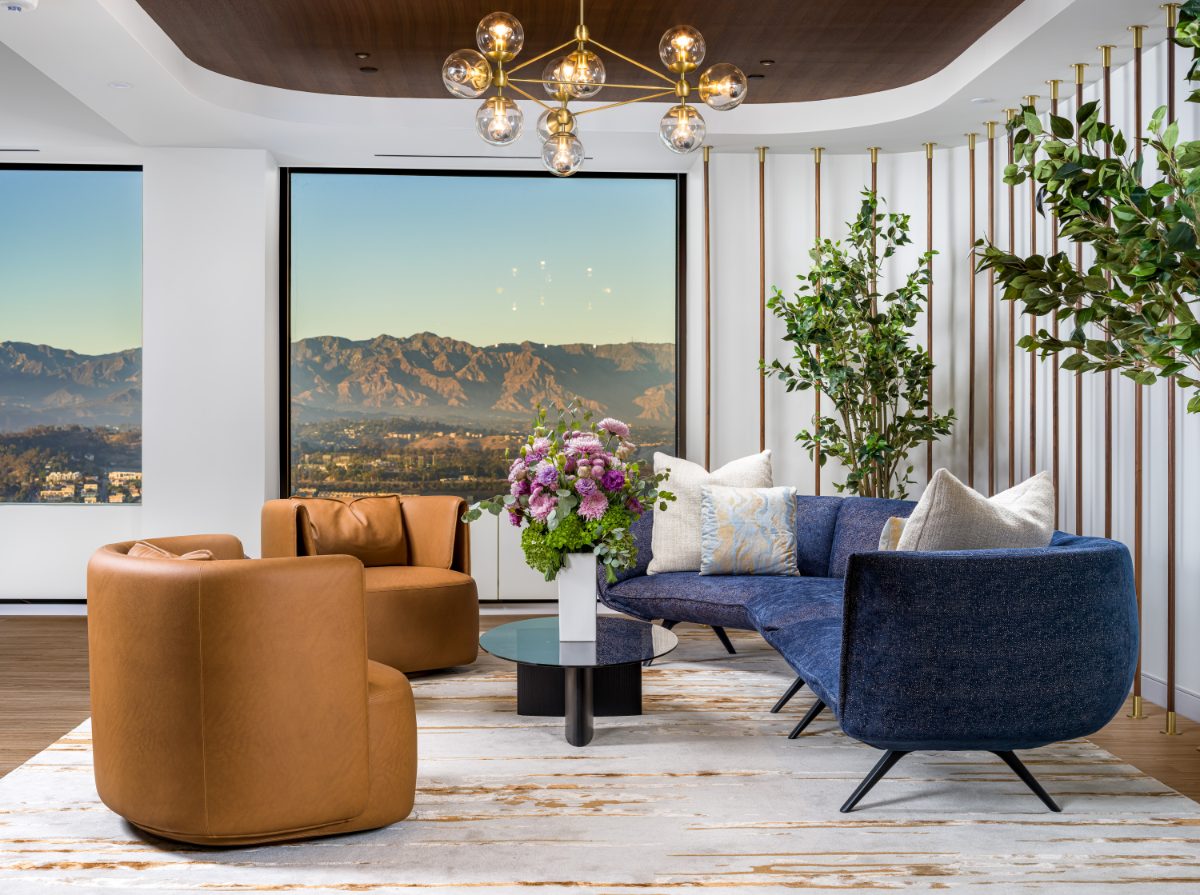 Golden-hour reception lounge at JAMS Los Angeles — navy chesterfield, cognac swivel chairs, brass globe chandelier, walnut ceiling, and hills through the window.