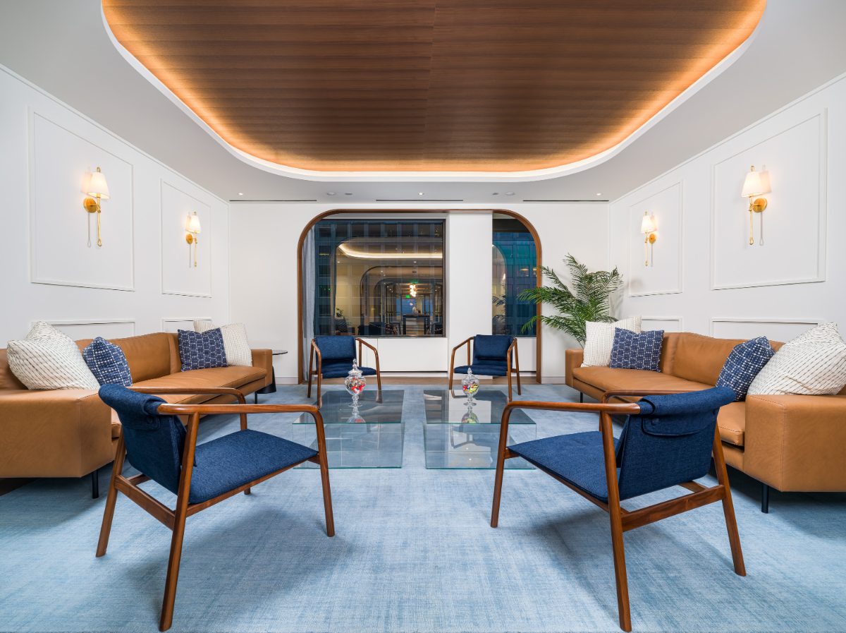 JAMS LA caucus lounge at blue hour — twin cognac sofas, blue mid-century chairs, brass sconces, walnut ceiling oval, neighboring tower lights through arched window.