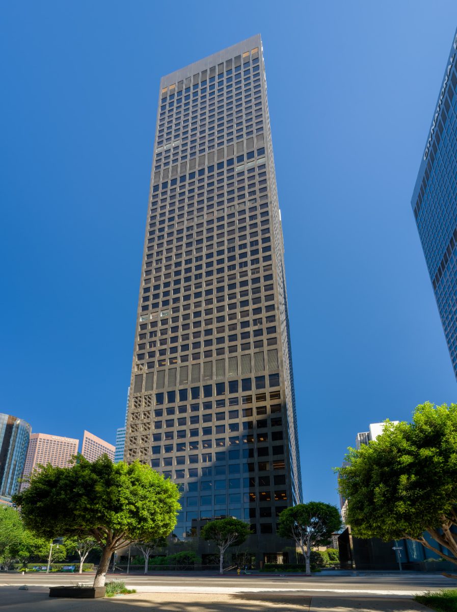 City National Plaza tower from across Flower Street — straight-on vertical exterior of JAMS LA's office building, framing ficus trees, midday sky.