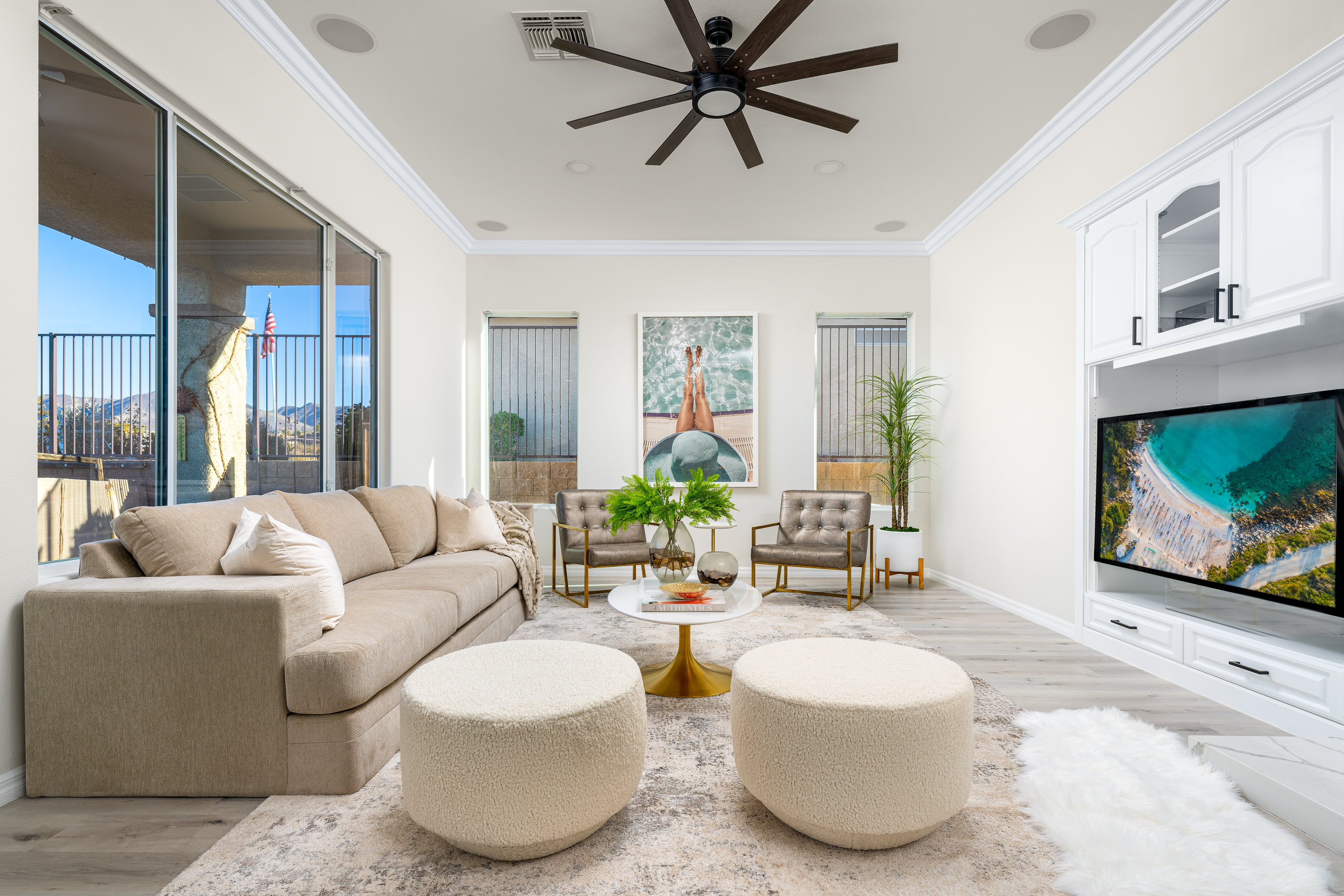 Living Room at a Stately casual coastal elegance home in Las Vegas (1952 OSS) — beige sectional, dark wood ceiling fan, gold-leg side tables, bouclé poufs.