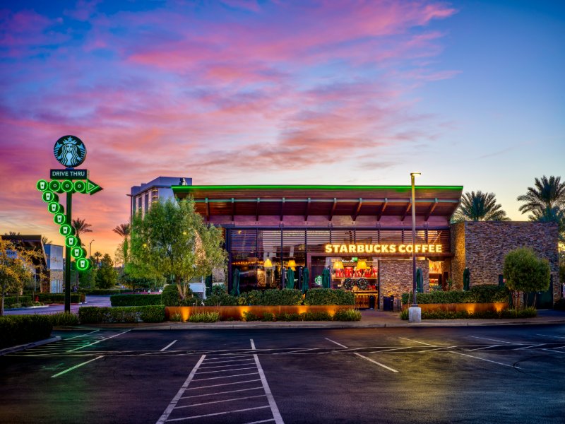 Starbucks drive-thru on Blue Diamond Road, Las Vegas at sunrise — pink-magenta sky, green neon Siren signage, warm interior lamps, wood-and-stone exterior, palm trees.