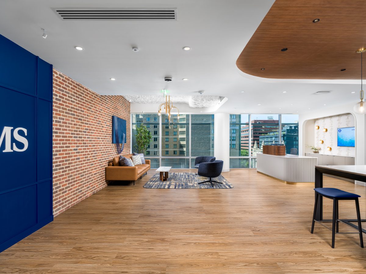 JAMS DC reception at 1050 K Street NW — cobalt brand wall, brick accent wall with tan leather sofa, central seating under brass chandelier, curved reception desk.