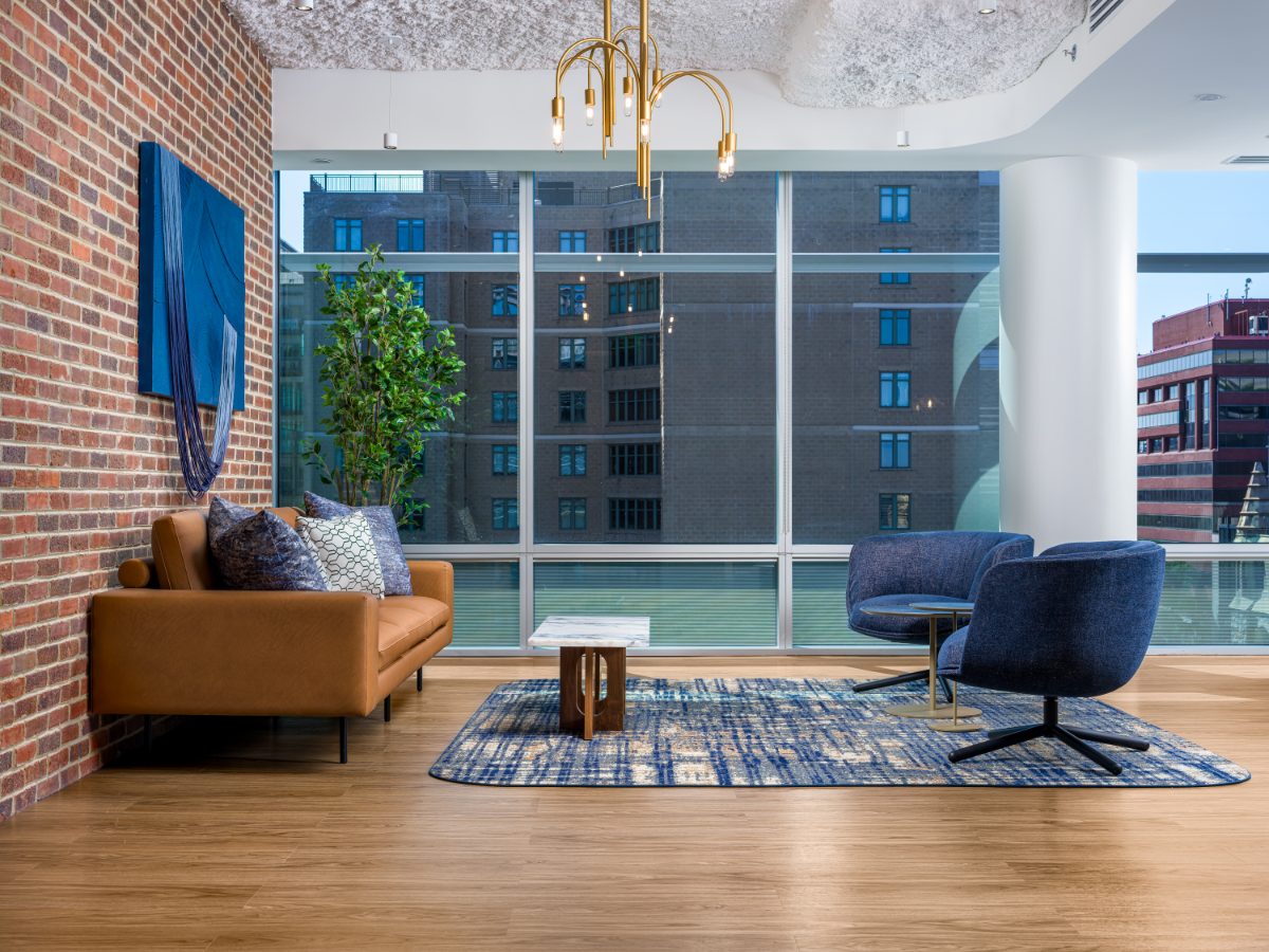 JAMS Washington DC reception waiting area — tan leather sofa, cobalt swivel chairs, exposed brick wall, brass chandelier, and floor-to-ceiling city view window.