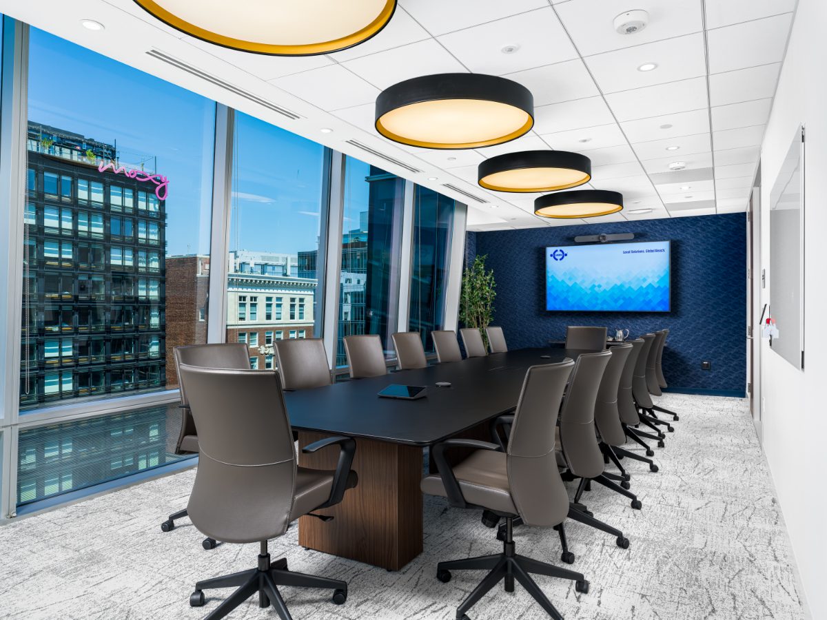 Large boardroom at JAMS Washington, D.C. — long black table, fourteen grey leather chairs, three drum pendants, navy wall with JAMS TV, and city-view windows.