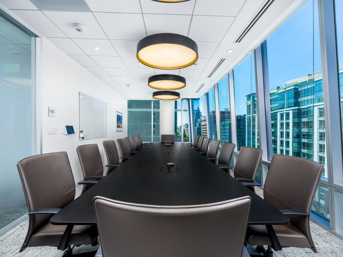 JAMS Washington, D.C. boardroom from the head of the table — long black table, fourteen grey leather chairs, drum pendants in perspective, wall-of-glass city view.