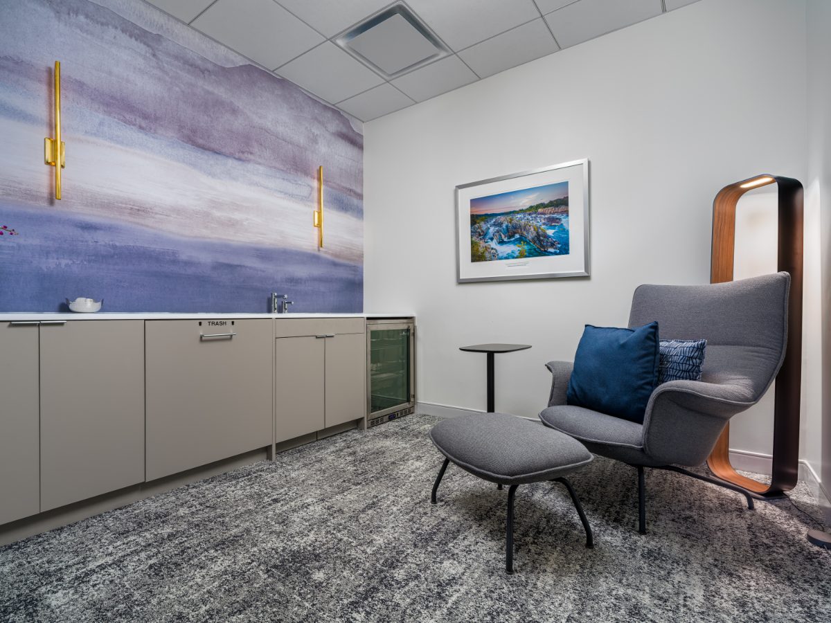 Wellness Center at JAMS Washington, D.C. — purple watercolor mural with brass sconces, grey lounge chair and ottoman, framed Great Falls print, and a kitchenette bar.