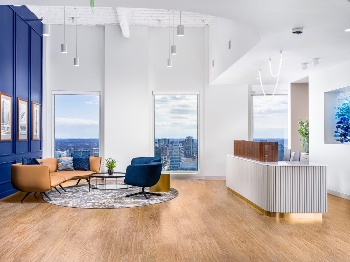 Wide-angle reception at JAMS Detroit — cobalt panel wall, cognac sofa, cobalt chairs, fluted reception desk with walnut top, and downtown skyline windows.
