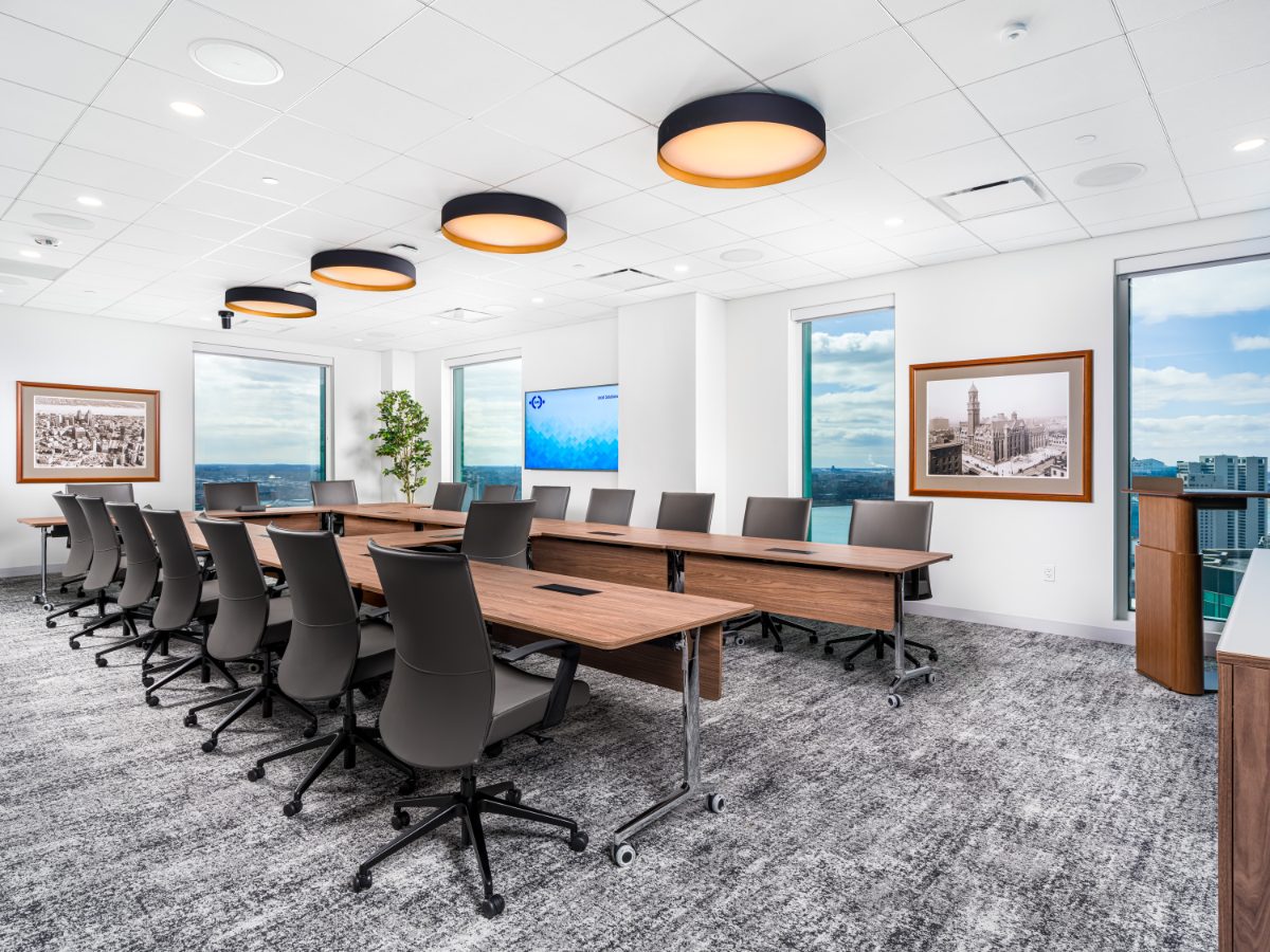 Grand boardroom at JAMS Detroit — U-shape of walnut tables, twenty grey leather chairs, four drum pendants, framed historic Detroit photos, and river-view windows.
