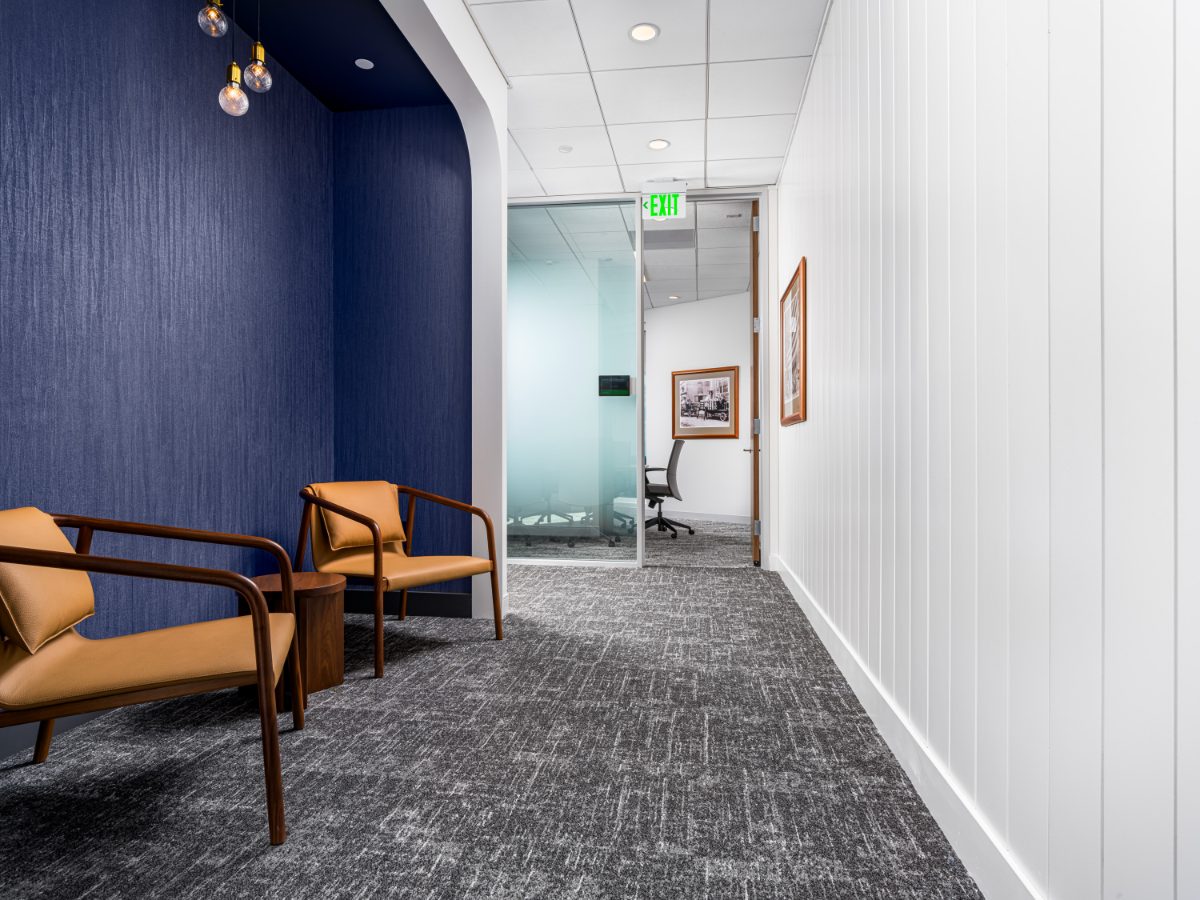 JAMS Detroit hallway lounge — navy alcove with Edison pendants and two cognac leather chairs, opening into a white vertical-board corridor with historic Detroit prints.