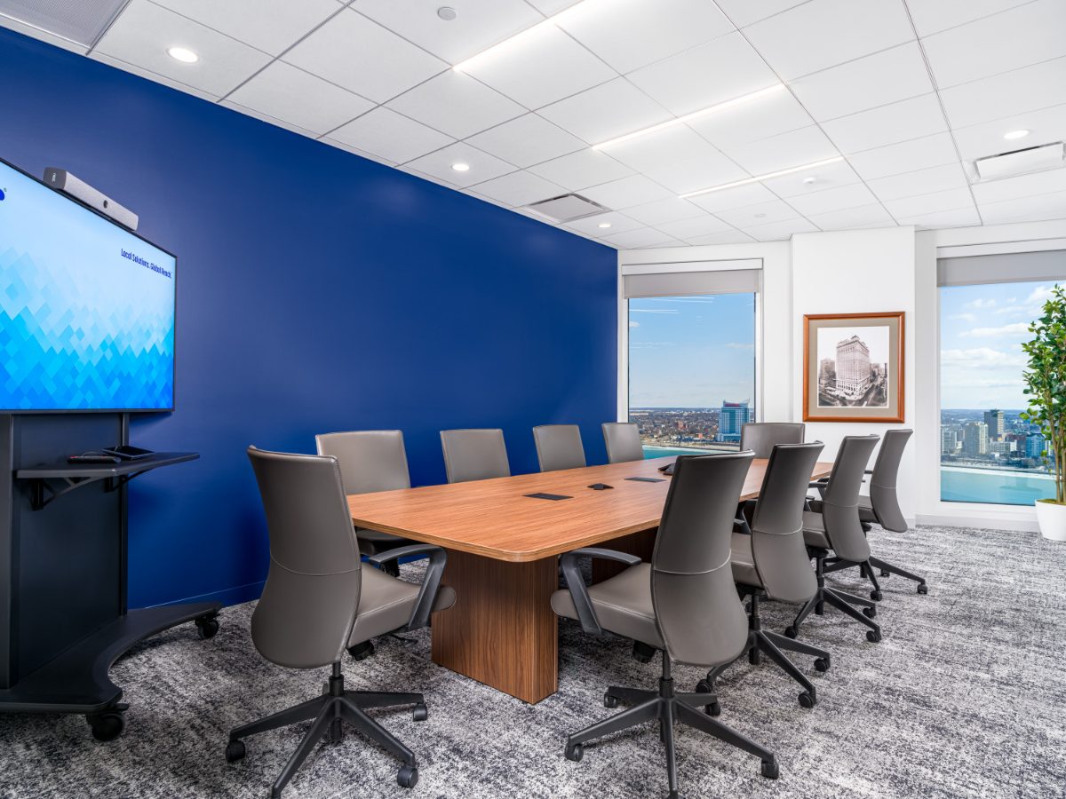Cobalt conference room at JAMS Detroit — walnut table, grey leather chairs, deep cobalt wall, mobile JAMS display, framed historic Detroit print, and river-view windows.