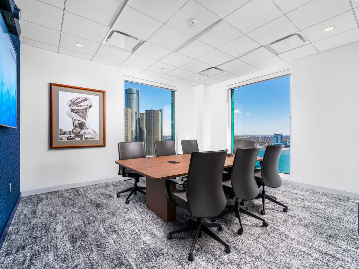 Corner conference room at JAMS Detroit — walnut table, six grey chairs, framed Vernor's print, and corner windows onto the GM Renaissance Center and Detroit River.