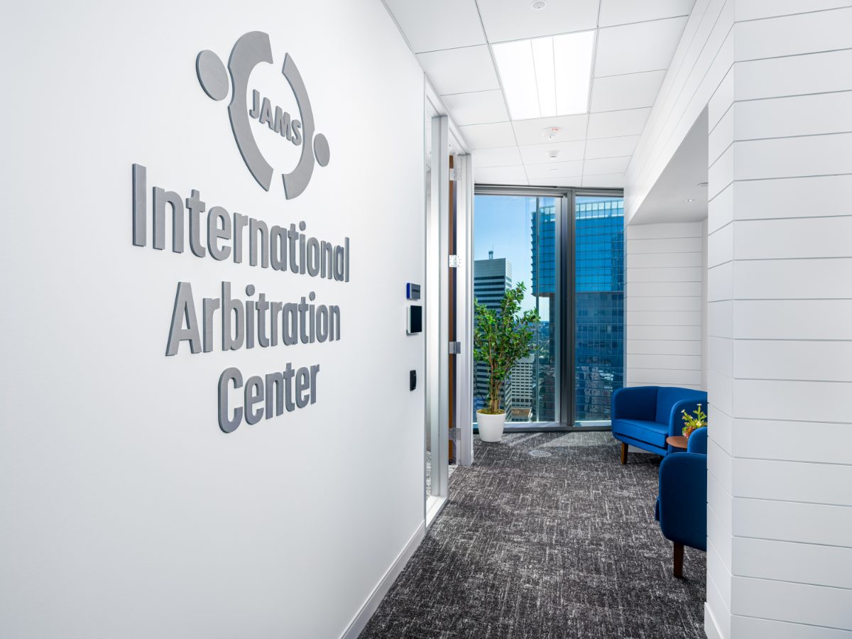 JAMS International Arbitration Center brushed-metal signage on a white wall at JAMS Houston, with a small lounge and skyline window beyond the doorway.
