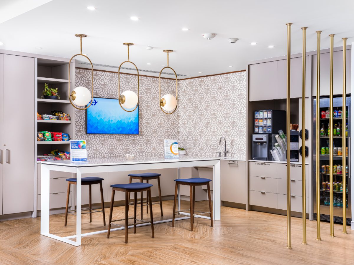 AMS Miami hospitality cafe on the 36th floor of One Biscayne Tower — brass-pendant counter island, blue-leather stools, snack shelves, stocked beverage cooler.