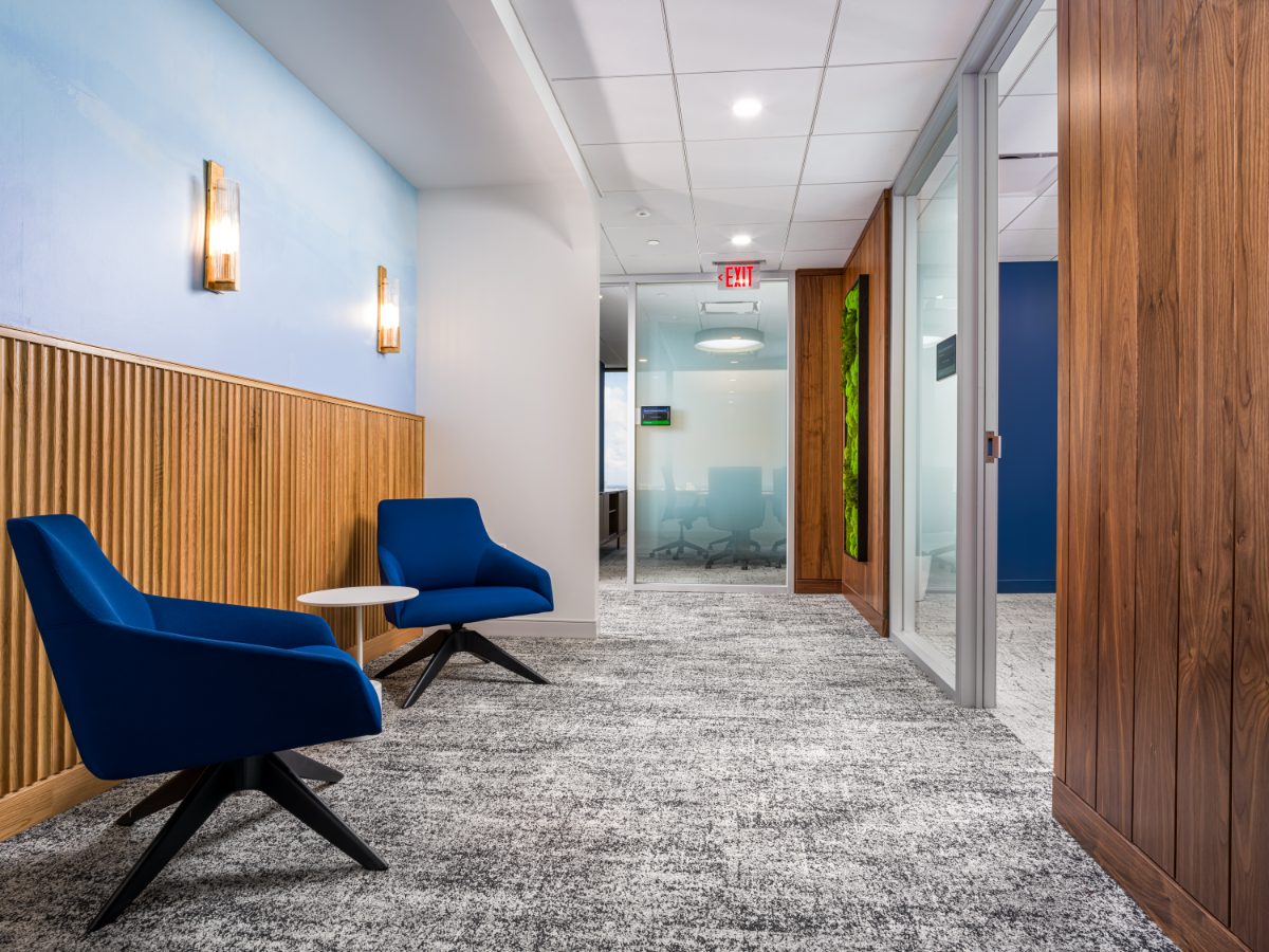 JAMS Miami hallway lounge between hearing rooms — two royal blue chairs, brass sconces, walnut wainscoting, and a moss wall art panel at the corridor end.