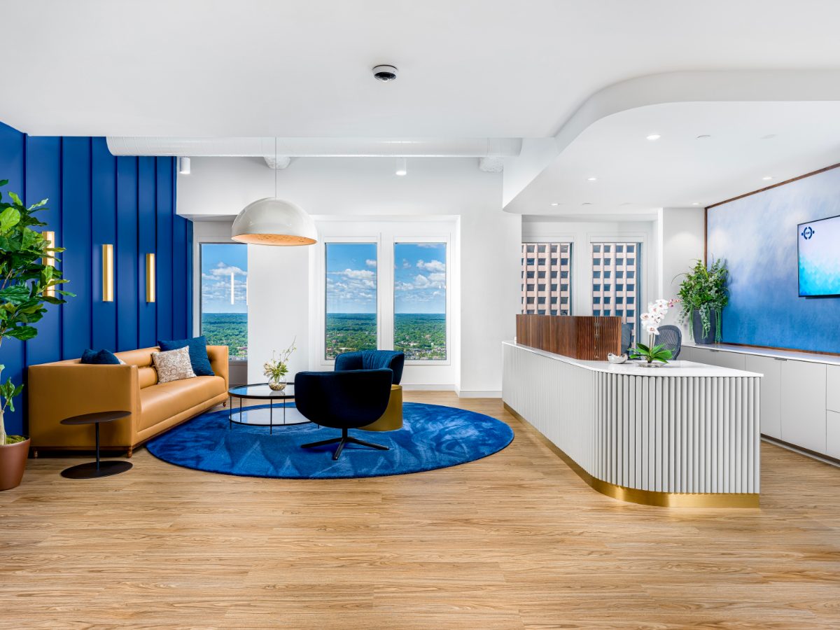 JAMS Minneapolis reception — fluted white desk with walnut panels, cobalt board-and-batten wall, cognac sofa, sweeping tree-canopy view through the windows.