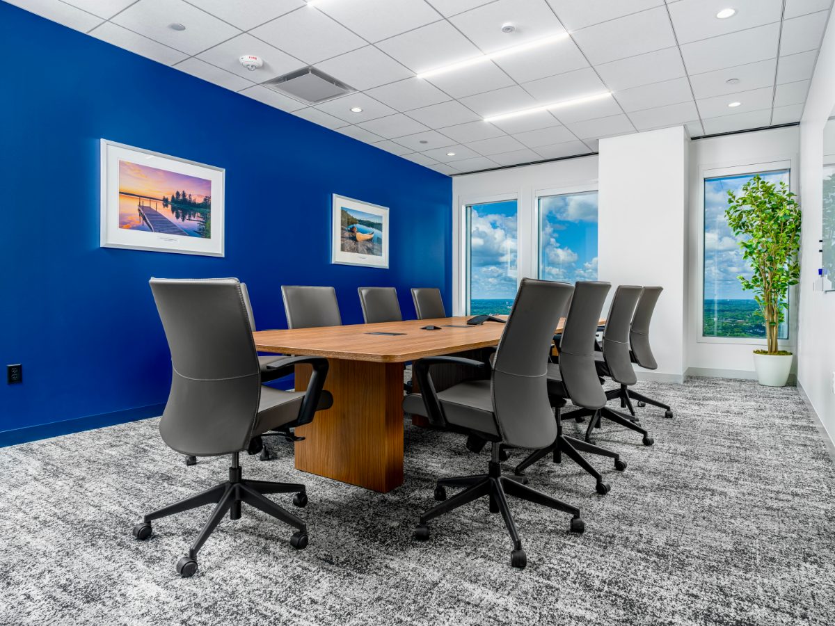 JAMS MN smaller conference room — honey-wood table on plinth, grey leather chairs, cobalt feature wall with framed Minnesota lake prints, suburban tree-canopy view.