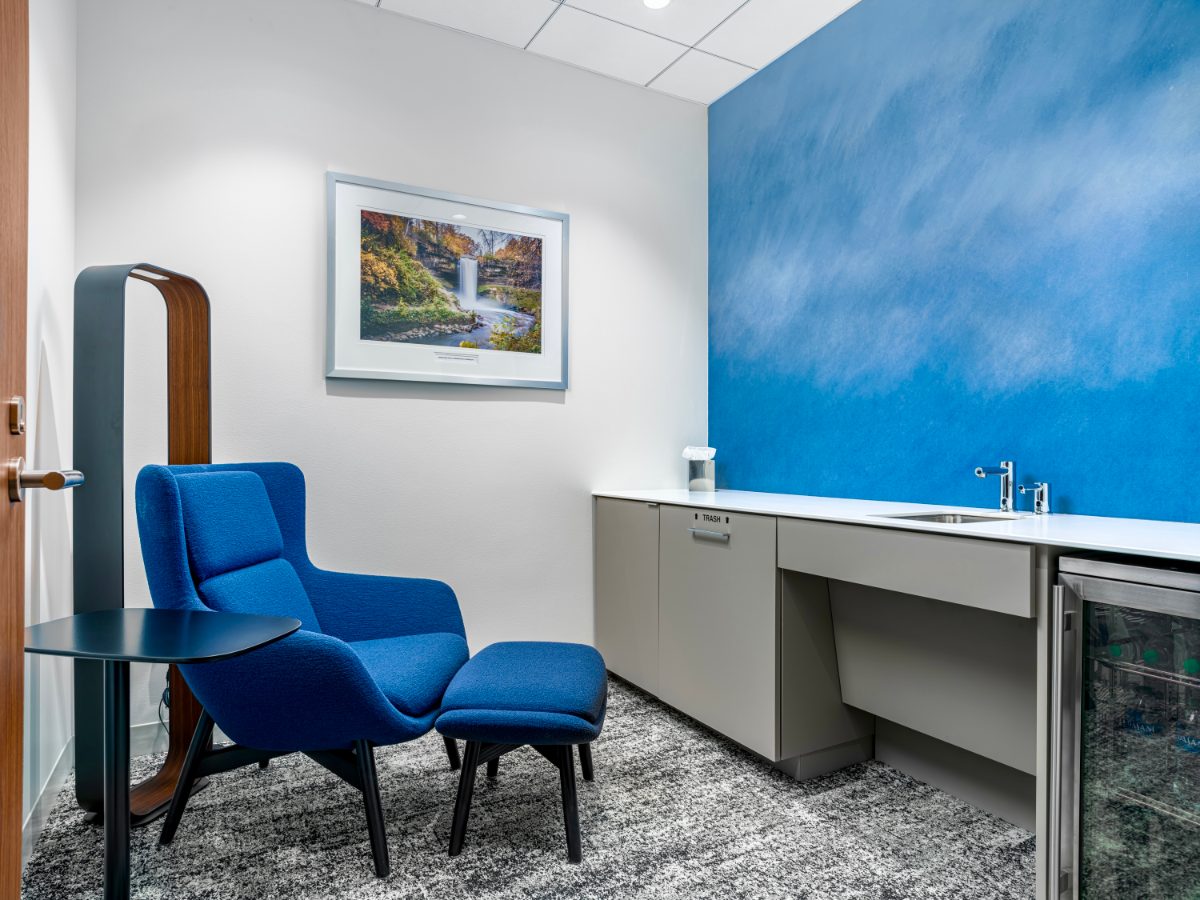 JAMS MN wellness room — cobalt swivel chair and ottoman, full-length mirror, framed Minnehaha Falls photo, cobalt-cloud painted feature wall, sink and beverage cooler.