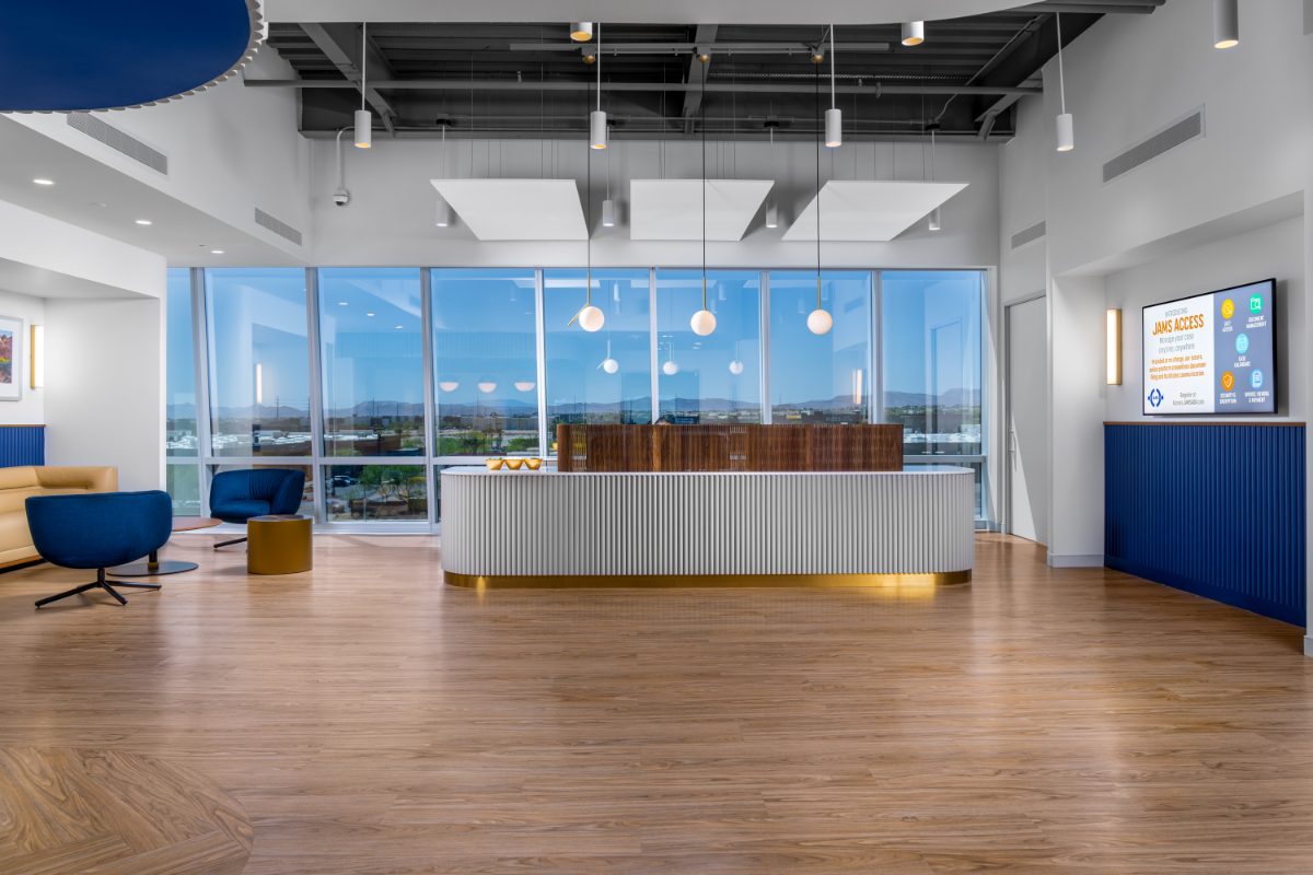 JAMS Las Vegas reception — fluted white desk with walnut panels, exposed industrial ceiling, globe pendants, cobalt swivel chairs, picture-window valley view.