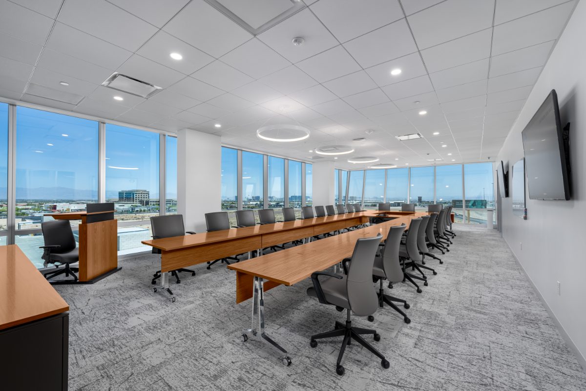 JAMS LV mega boardroom — honey-wood U-configured table with grey leather chairs, halo ring pendants, wraparound windows framing the Las Vegas Strip on the horizon.