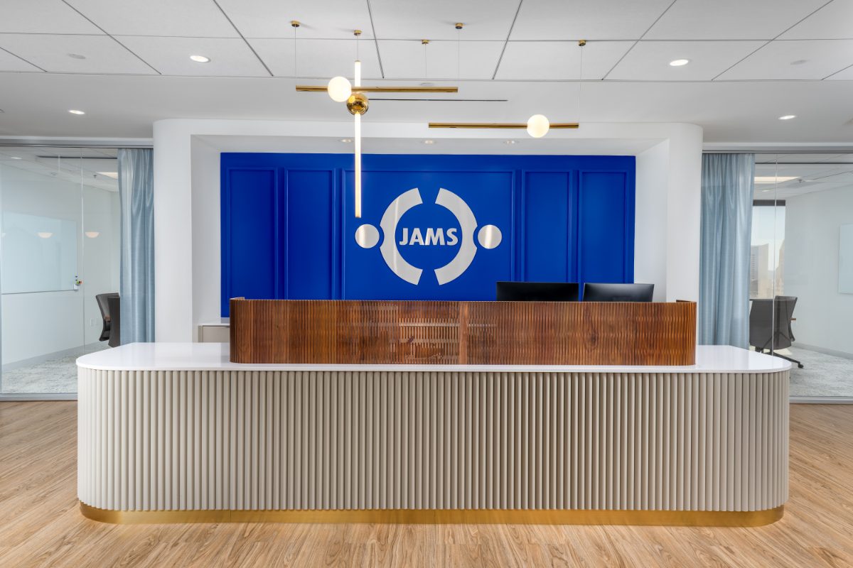 JAMS San Diego reception, Symphony Towers — oval fluted desk with brass kick, walnut slat screen, cobalt feature wall with brushed JAMS emblem, twin brass globe pendants.
