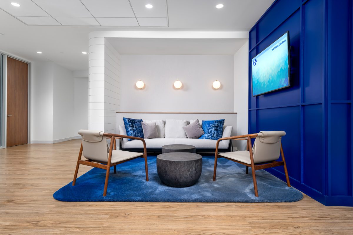JAMS San Diego reception lounge — walnut lounge chairs with cream sling cushions, tufted sofa with indigo pillows, charred-wood drums on cobalt rug, brass sconces.