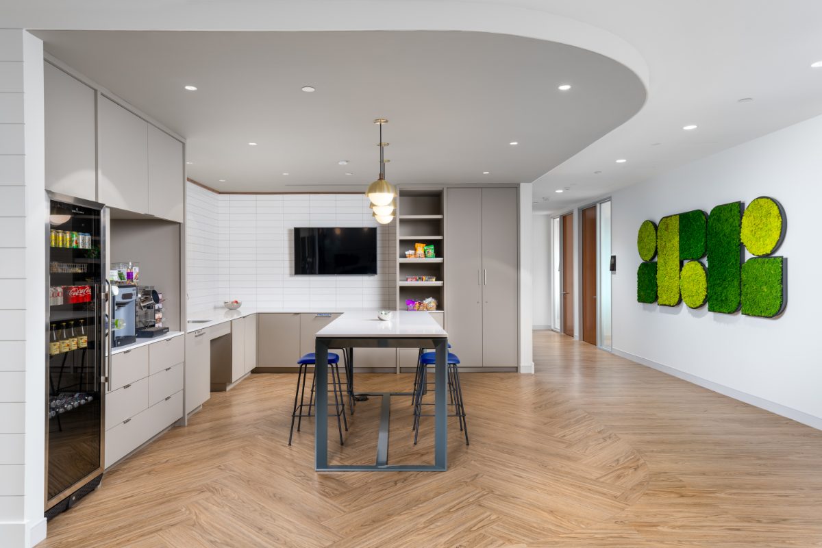 JAMS San Diego Café — curved soffit over quartz high-top with cobalt stools, glass-front fridge, espresso bar, JAMS moss-emblem wall sculpture, chevron oak floor.