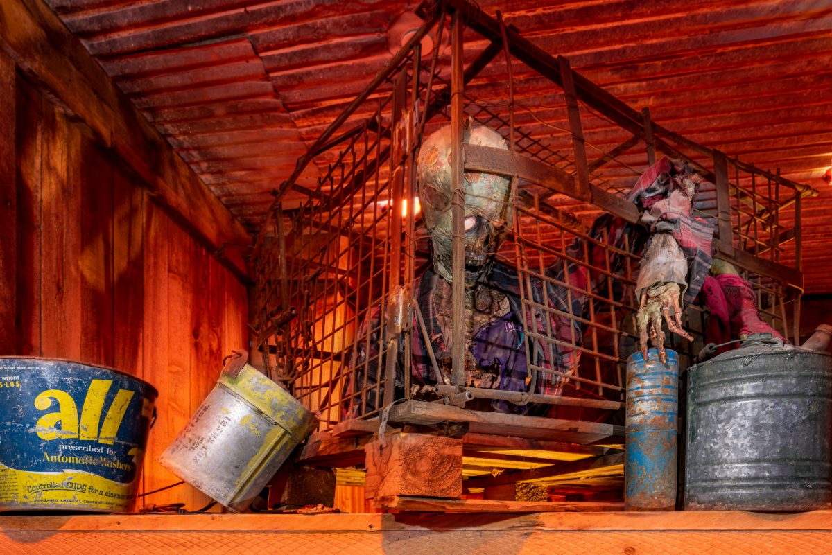 Detail shot inside the Zombie Outbreak escape room at PanIQ Las Vegas — a rusted iron-mesh cage in the loft of the barn set holding a desiccated zombie figure in ratty clothing, wi