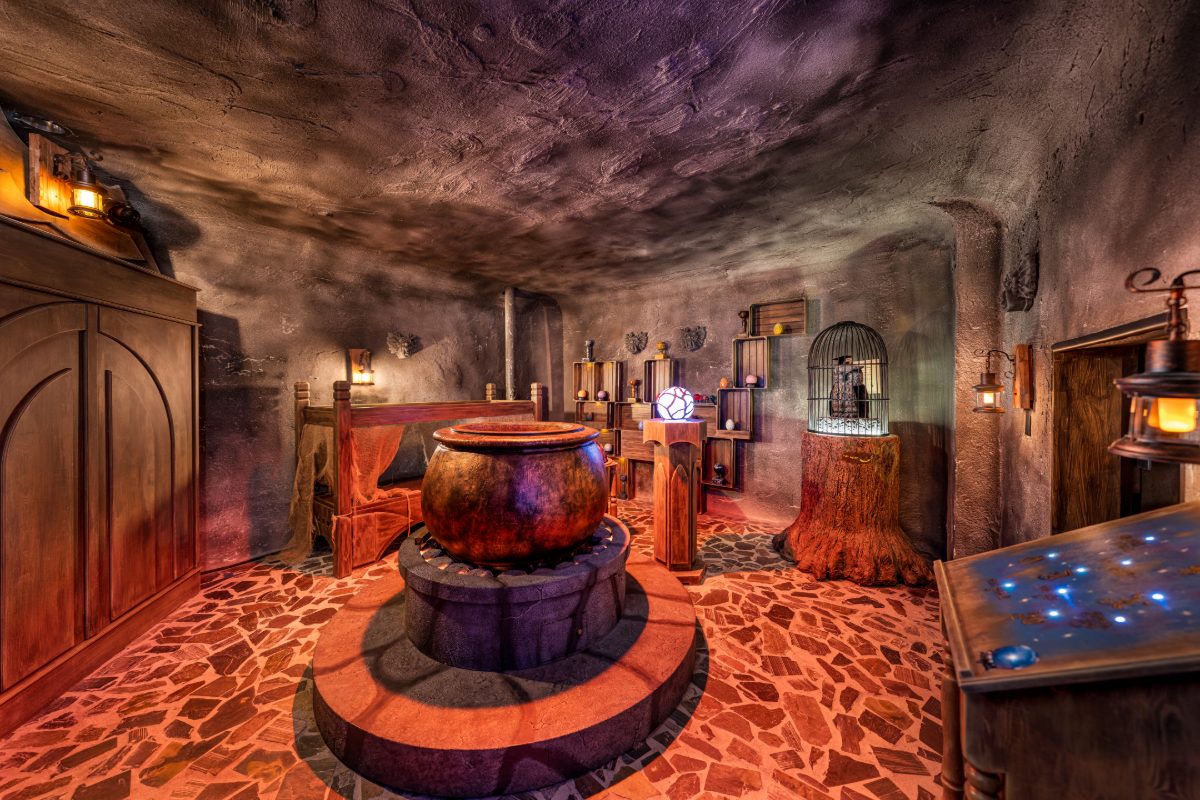 Interior of the alchemy chamber inside the Wizard Trials escape room at PanIQ Las Vegas — a sculpted cave-like room with curved earthen walls and ceiling, a massive iron cauldron o