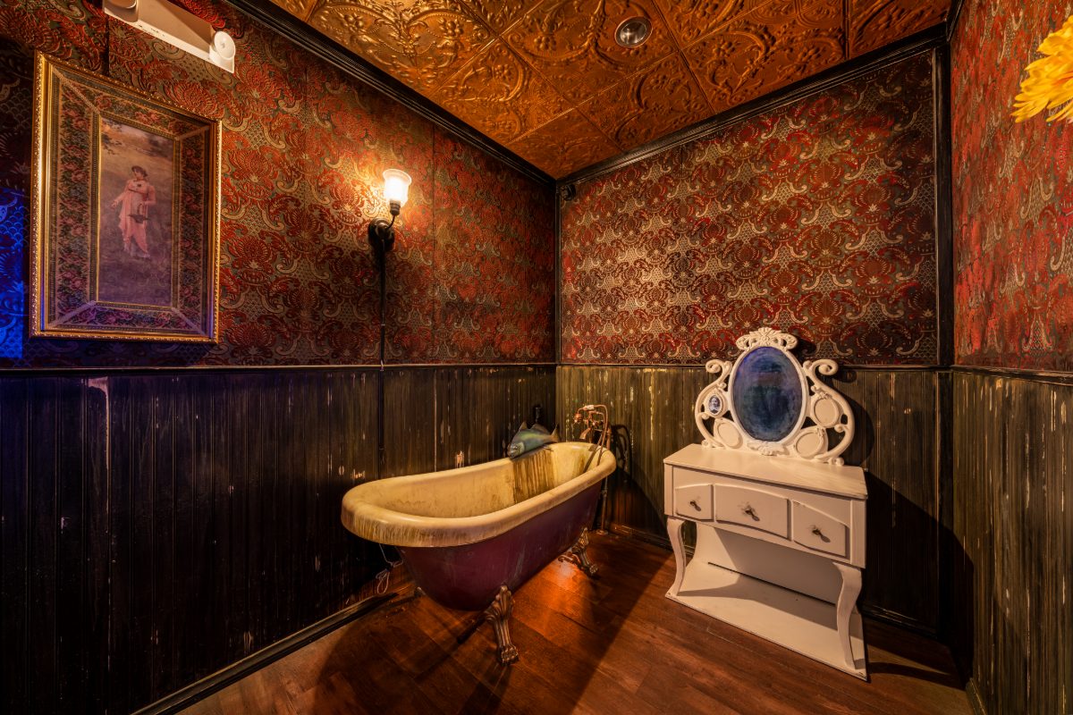 A bath alcove inside The Haunted Manor escape room at PanIQ Las Vegas — embossed copper pressed-tin ceiling, crimson and gold Victorian damask wallpaper, distressed black beadboard
