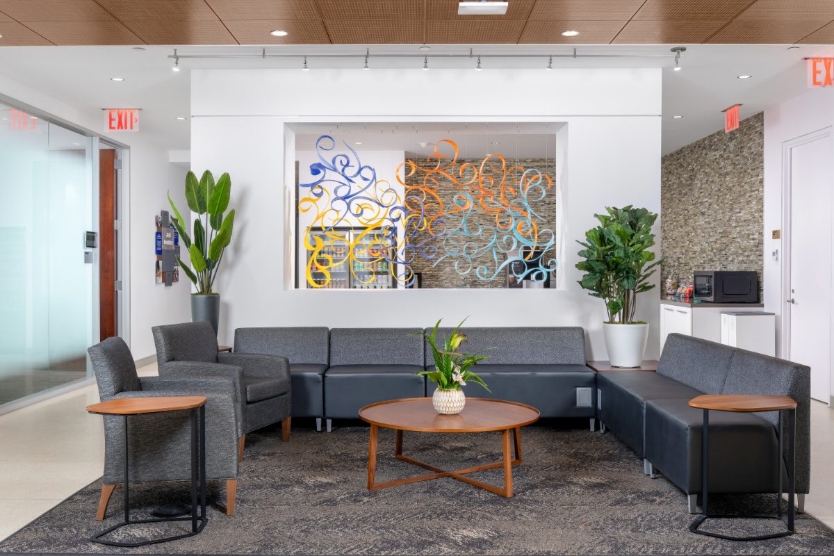 JAMS NYC reception lounge — grey sectional around walnut coffee table, colorful abstract sculpture over pass-through to JAMS Café, tall plants flanking.