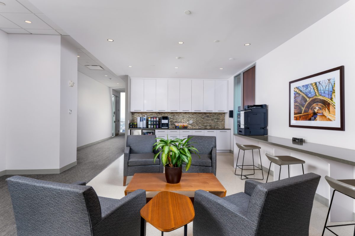 JAMS NY small café lounge — grey armchairs around warm wood coffee table, kitchenette with coffee station, work bar with printer, framed City Hall Station print.